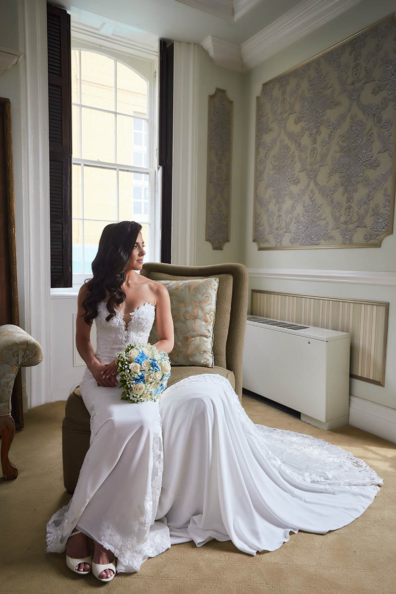 Bride sitting on chair in wedding dress looking out the window and holding flowers