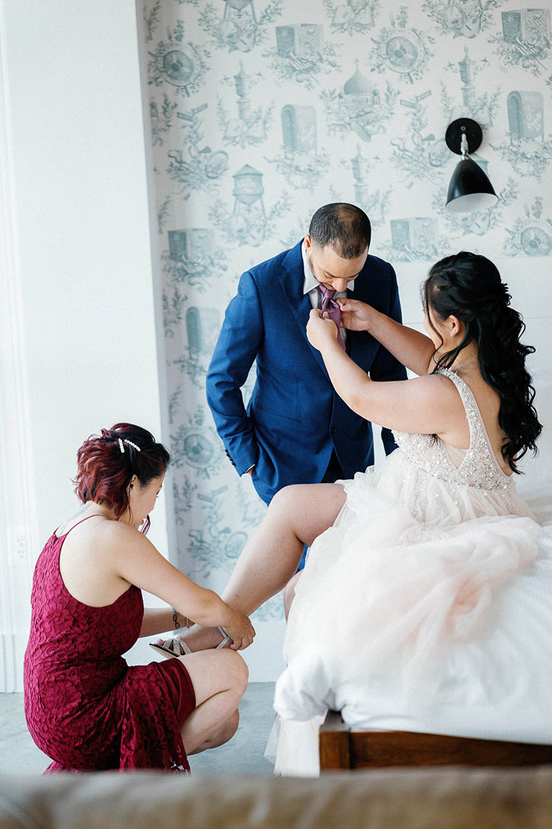 Maid of honor helping bride put the shoes on and bride helping groom fix his tie